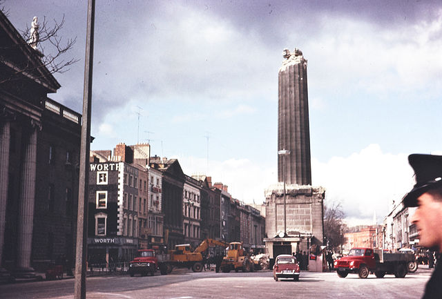 A photo of a street with a large column that is missing its top, with cars and people on the street and historic buildings lining the road 