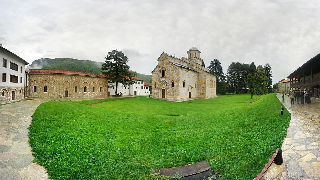 a green lawn with a church and other historic buildings on it, people stand on the paving stones and there is a hill in the background