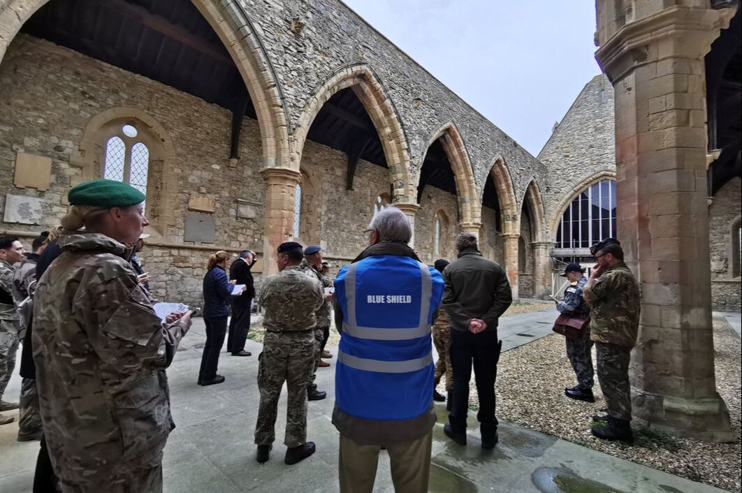 A man with a high vis vest saying “Blue Shield” stands amongst a group of people in military uniform in the middle of an old stone building with archways and pillars