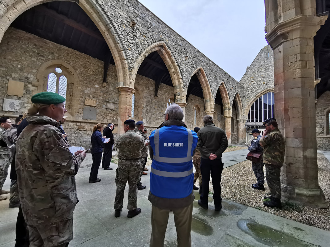 A man with a high vis vest saying “Blue Shield” stands amongst a group of people in military uniform in the middle of an old stone building with archways and pillars