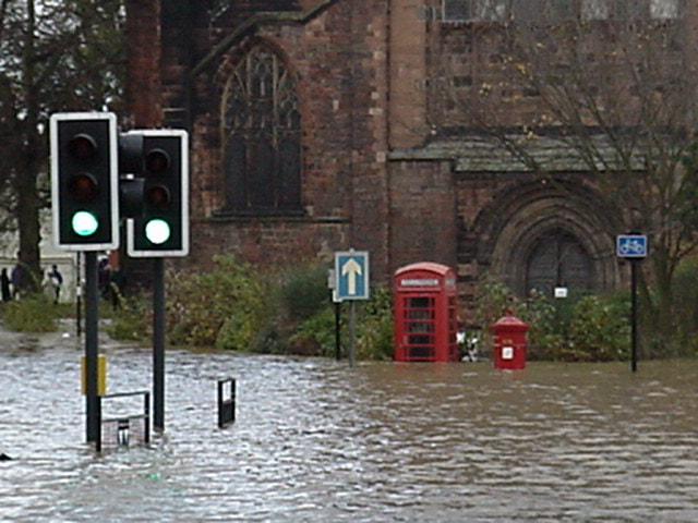 A street that has flooded with the water going halfway up the traffic lights and a historic red telephone and red post box and historic church in the background 