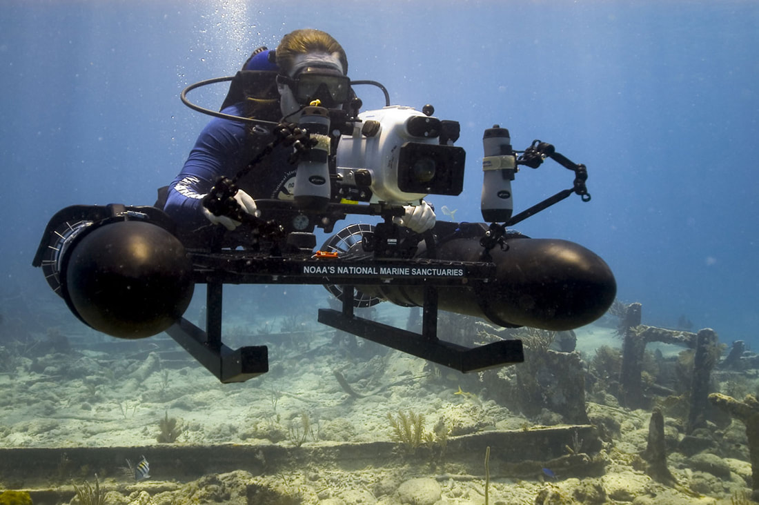A man scuba diving with a motorised camera on the sea floor with some ruins in the sand